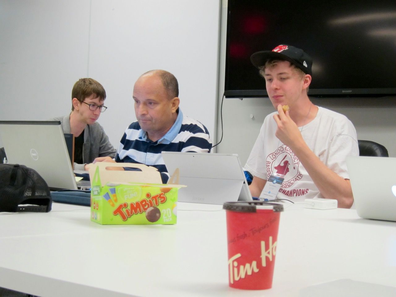 HelloNewman eating a Timbit while nafes & Cottser concentrate on their work. In the foreground is an iconic Tim Hortons coffee.